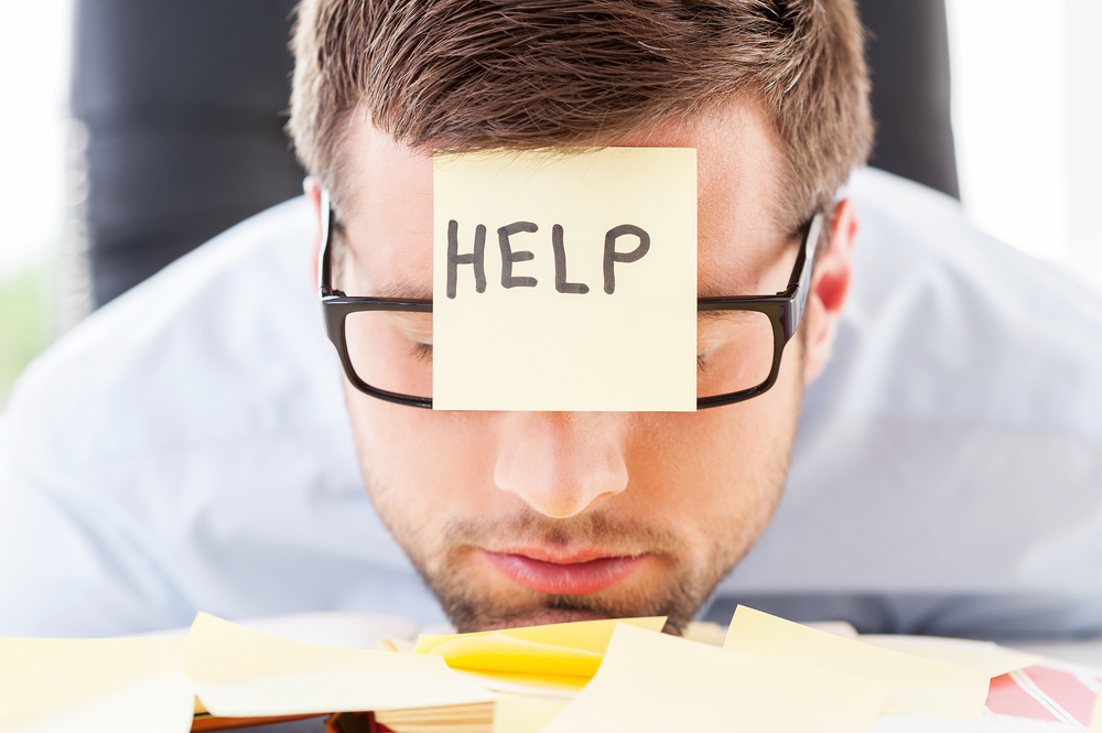 person with their chin on a desk and a post-it note on their forehead reading 'help'
