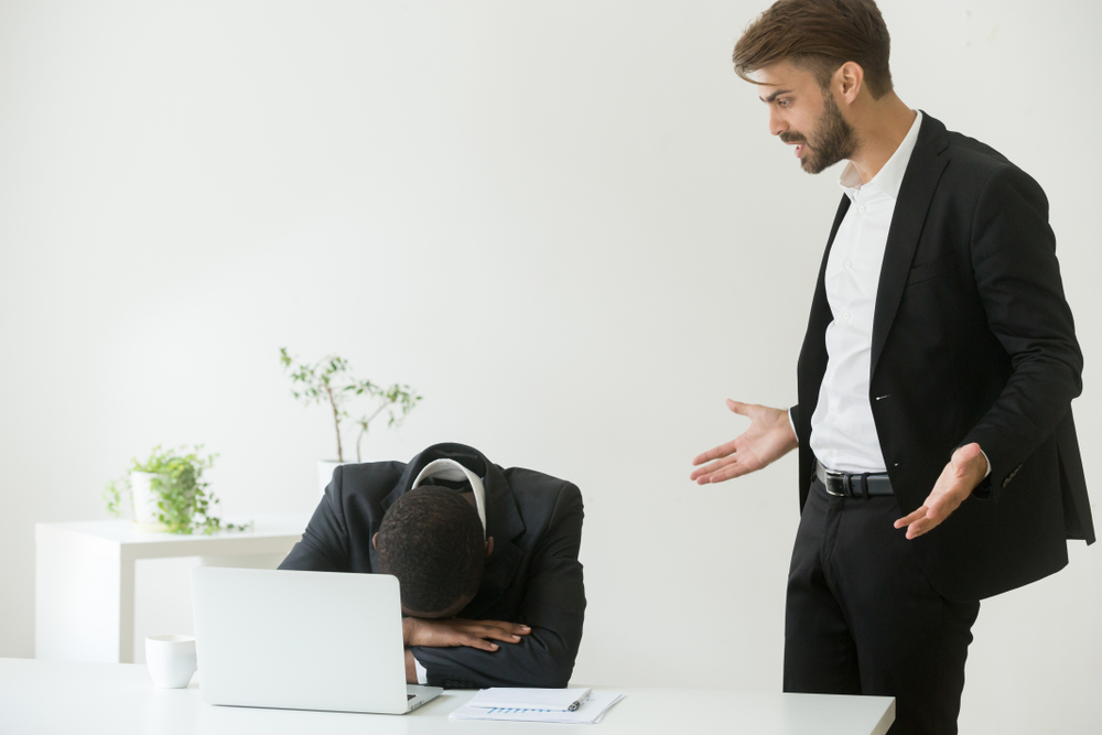 Employee with head down on desk while manager stands over looking frustrated